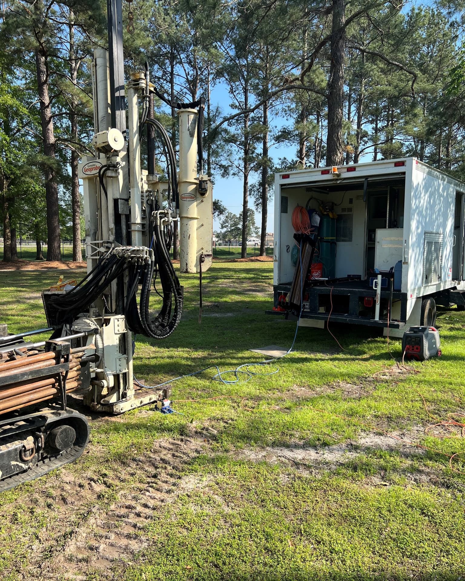 A direct-push platform set up on a grassy site under pines, with the data acquisition truck parked behind it, its rear doors open