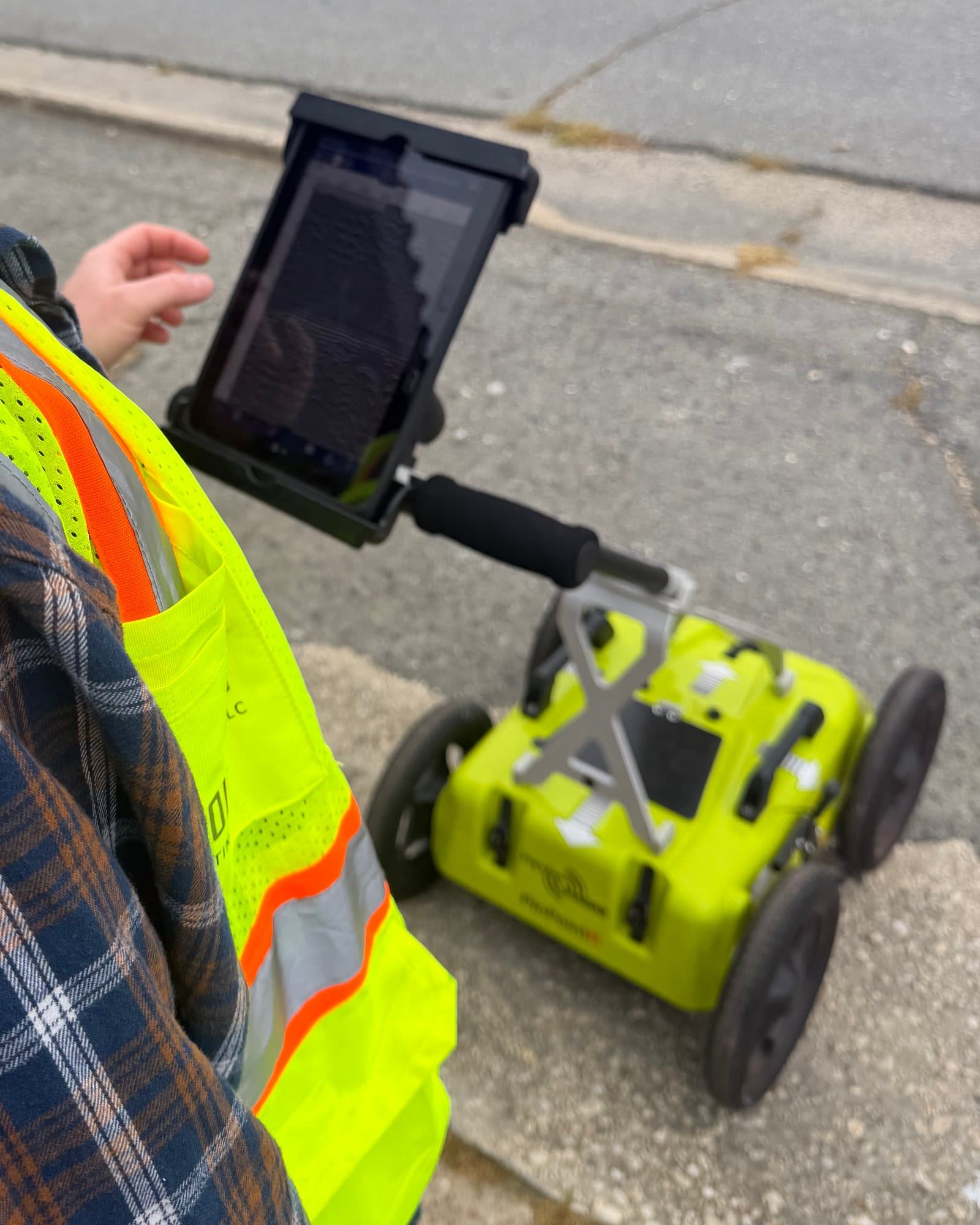 A technician in a high-visibility vest reads the radar on a tablet mounted to the handle of a ground-penetrating radar cart.